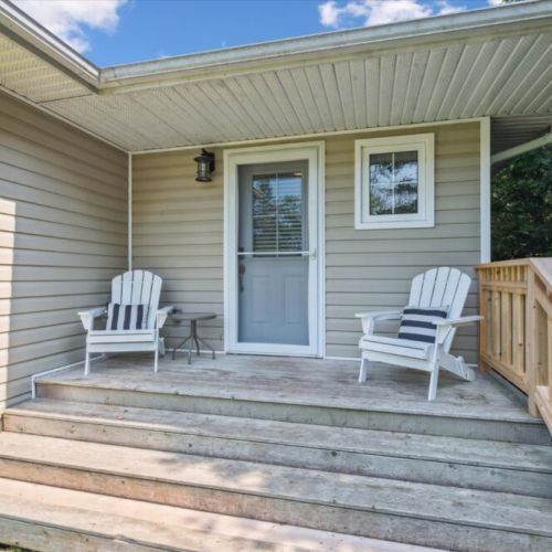 Welcoming front porch with two classic white Adirondack chairs, striped accent pillows, and a small side table — perfect for a quiet morning coffee or evening relaxation.