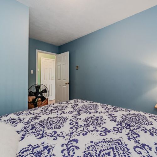 A simple yet elegant bedroom featuring tranquil blue tones and soft furnishings. The bed, adorned with patterned bedding and accent pillows, invites you to sink in and relax. The room includes a convenient fan for warm nights and ample closet space for your belongings, ensuring a clutter-free stay. Perfect for guests seeking calm and comfort.