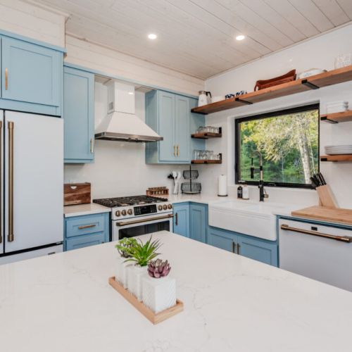 Bright and stylish vacation rental kitchen with open shelving, high-end appliances, and a serene forest view—perfect for travellers seeking a cozy culinary escape near nature and the lake.