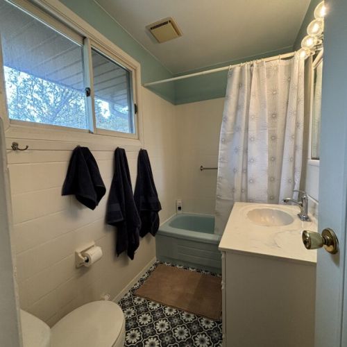 Crisp Bathroom Elegance: The bathroom pairs classic fixtures with crisp white and soft blue tones to create a refreshing space for guests to refresh and rejuvenate. Natural light filters through the window, highlighting the clean lines and inviting warmth of this well-maintained bathroom.