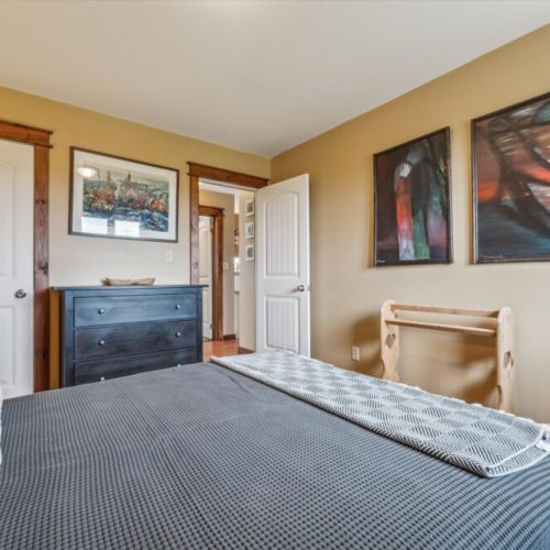 Main-level queen bedroom with rich wood floors, a sturdy three-drawer dresser, soft lighting, and local artwork adding a personal touch.